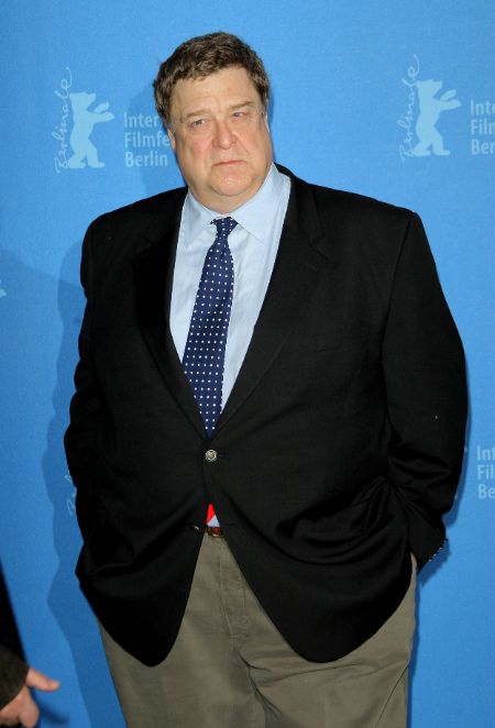 John Goodman in a black coat and sky-blue shirt poses for a picture.
